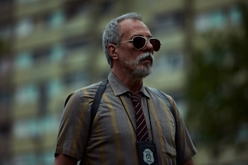 Un hombre con barba gris, gafas de sol, camisa a rayas y corbata, lleva una credencial de la Policía Provincial. Fondo de edificios borroso