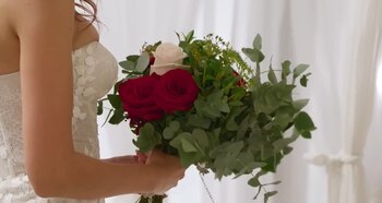 Primer plano de una persona con un vestido de novia blanco de encaje, sosteniendo un ramo con tres rosas rojas, una rosa blanca y hojas verdes