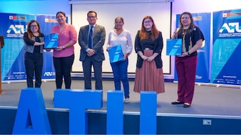 Seis personas, cinco mujeres y un hombre, posan en un escenario con el logotipo de ATU en el fondo y en bolsas. Hay letras grandes "ATU" en el suelo