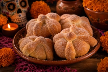 Panes cubiertos de azúcar en un plato de barro, rodeados por flores de cempasúchil anaranjadas, una vela encendida y una calavera decorada.
