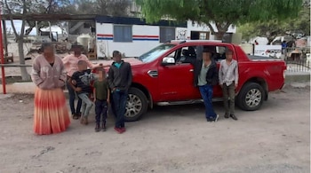 Grupo de siete personas, incluyendo adultos y niños, de pie junto a una camioneta pick-up roja. Al fondo, edificaciones modestas y árboles secos