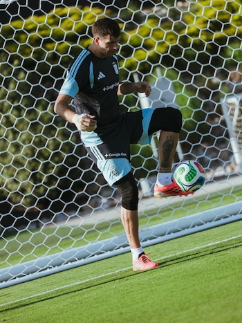 Dibu Martínez, con uniforme de Argentina, controla un balón de fútbol con su rodilla izquierda en un campo verde. Detrás, una portería con red hexagonal visible