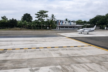Avioneta blanca de dos hélices en pista de aeropuerto. Edificio con fachada geométrica de colores y árboles tropicales al fondo.
