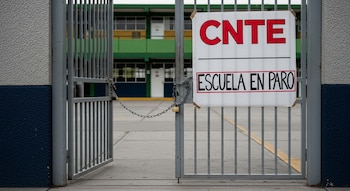 Reja de una escuela primaria pública mexicana cerrada con candado y cadenas. Un cartel de la CNTE con 'Escuela en paro' cuelga. Se observa el patio interior vacío.