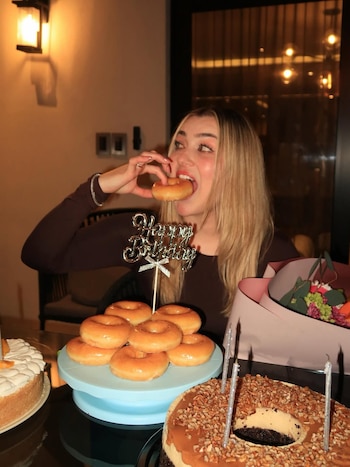 Melenie Carmona, una joven rubia, muerde una dona glaseada en una fiesta de cumpleaños. Detrás hay una torre de donas, dos pasteles y flores