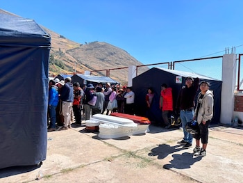 Grupo de personas de pie al aire libre, dos tiendas de campaña azules, tres ataúdes y un fondo de montañas bajo cielo azul