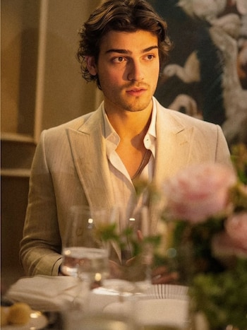 Luca Bertoldi Menem, con cabello castaño ondulado, barba incipiente y traje claro, sentado en una mesa con copas y flores borrosas