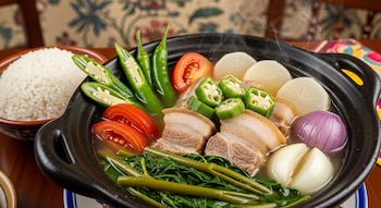 Primer plano de Sinigang de cerdo filipino en cazuela negra, con carne, tomates, okra, rábano blanco, espinaca de agua y cebolla. Arroz blanco al lado.