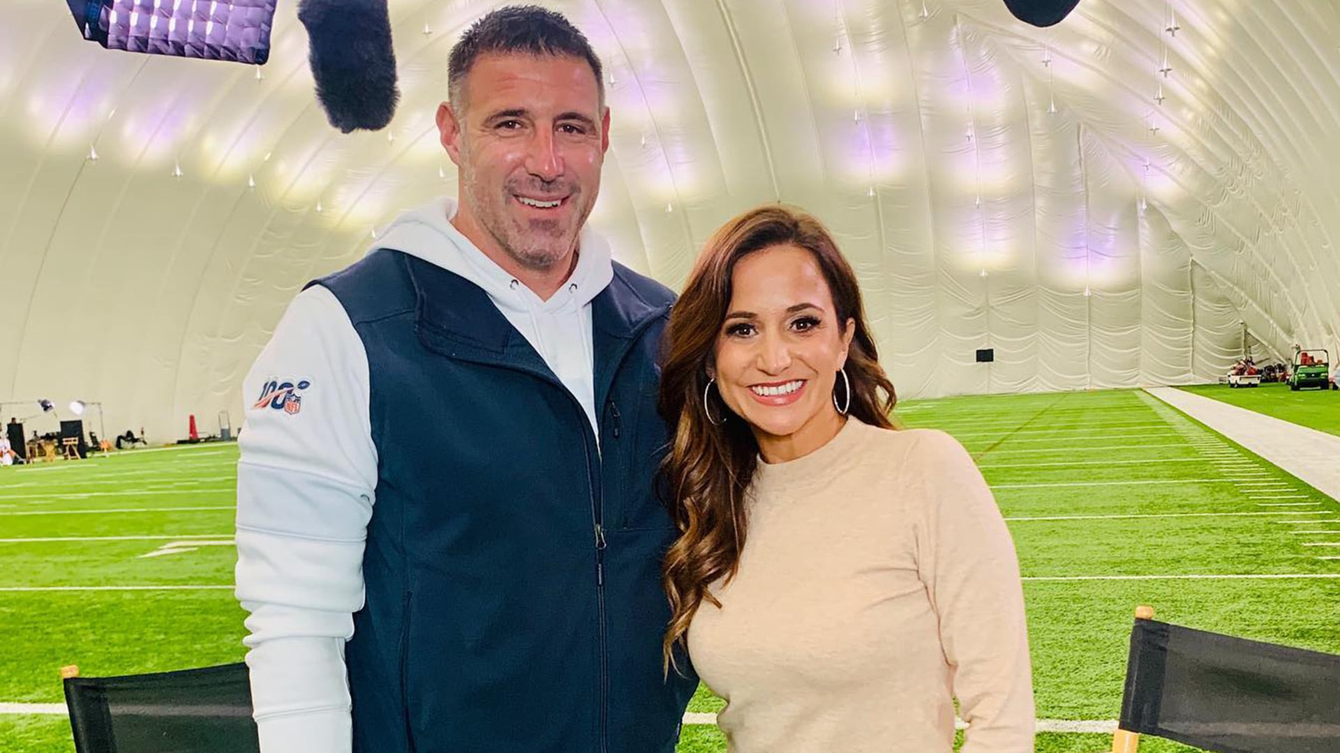 La periodista deportiva Dianna Russini posa sonriente junto al entrenador Mike Vrabel en una instalación deportiva cubierta para una entrevista antes de la temporada