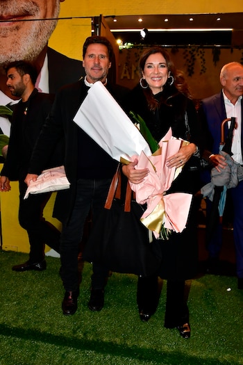 Un hombre y una mujer sonrientes, vestidos de oscuro, sostienen ramos de flores y regalos envueltos. Están de pie sobre césped artificial