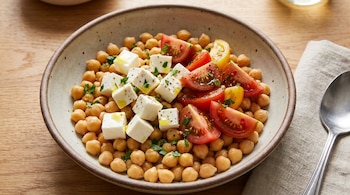 Un bowl con garbanzos cocidos, cubos de queso blanco, rodajas de tomate rojo y amarillo, perejil picado, aceite de oliva, y una cuchara en un paño sobre madera.