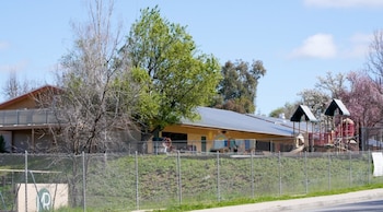 Una escuela de un piso con techo gris y paredes beige, detrás de una valla de tela metálica. Se ven árboles verdes y florecidos, y un área de juegos. Cielo azul