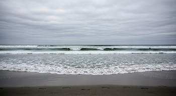 Playa de arena oscura con huellas y olas espumosas. Agua del mar oscura con dos siluetas en la distancia y un cielo completamente nublado.