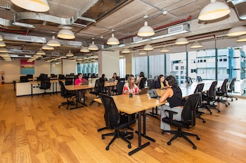 Amplia oficina moderna con piso de madera, mesas de madera y sillas negras. Varias mujeres usan computadoras portátiles. Ventanales y edificios exteriores
