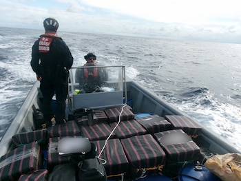 Dos guardacostas con cascos a bordo de una pequeña embarcación en el mar, rodeados de paquetes rectangulares apilados