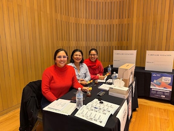 Tres mujeres sonrientes en una mesa con materiales electorales, incluyendo urnas de cartón y letreros de 'CABINA DE VOTACIÓN', en un centro de votación