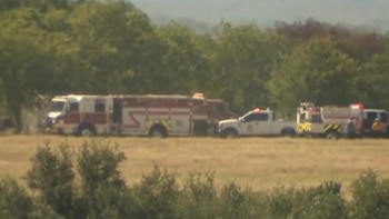Un camión de bomberos blanco y rojo, una camioneta blanca y un vehículo de emergencia más pequeño estacionados en un campo de hierba seca con árboles al fondo