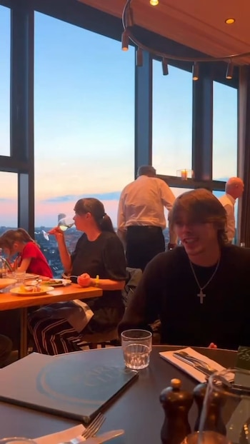Marta Fort y Felipe Fort cenando en un restaurante de altura con grandes ventanales, ofreciendo una vista panorámica de la ciudad al atardecer. Marta bebe de una copa, Felipe sonríe al frente
