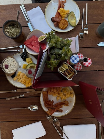 Mesa de madera con platos de desayuno, mate, huevos revueltos, tocino, aguacate, sandía, fruta de la pasión, avena, jaleas, cubiertos y servilletas