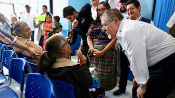 Un Presidente de barba gris, vestido con camisa blanca, conversa con una mujer sentada en una silla azul en una instalación de salud