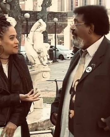 Una mujer con chaqueta negra habla frente a un hombre con traje, anteojos y barba en una plaza con una escultura al fondo