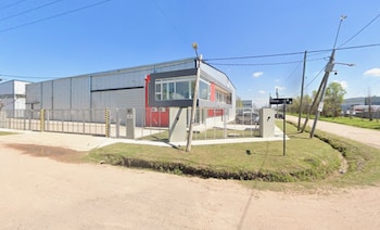 Edificio industrial de metal y fachada roja con reja frontal, acera de hormigón, calle de tierra y postes de luz bajo un cielo despejado