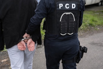 Vista trasera de un individuo esposado siendo escoltado por un oficial de policía con chaleco táctico que dice PCD.