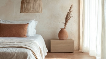 Vista parcial de un dormitorio con cama, almohada naranja, lámpara de mimbre, mesita de noche de madera, jarrón de barro con planta seca y cortinas blancas.