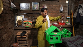 Hombre de mediana edad con gafas, dos casas para pájaros verdes, pegatinas de banderas de India, pared de ladrillo, estantería, fotos, premio, manta