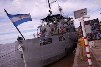 Buque militar gris con la bandera de Argentina en la popa, amarrado a un muelle junto a contenedores de carga y postes de señalización