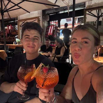 Un hombre sonriente y una mujer con labios fruncidos brindan con cócteles de color naranja en un bar por la noche