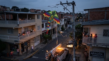 Calle de barrio al anochecer con personas en balcones y en la calle usando velas. Un camión de servicio y una cometa colorida enredada en los cables.