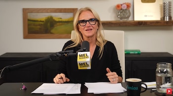Una mujer con gafas negras sentada frente a un micrófono con el logotipo de Mel Robbins, hojas de papel y una taza