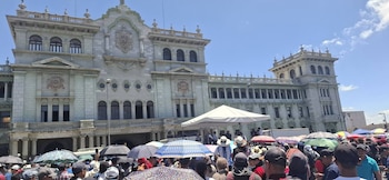 Multitud de personas con sombrillas frente al Palacio Nacional de la Cultura de Guatemala. Se observa un toldo blanco y el cielo azul