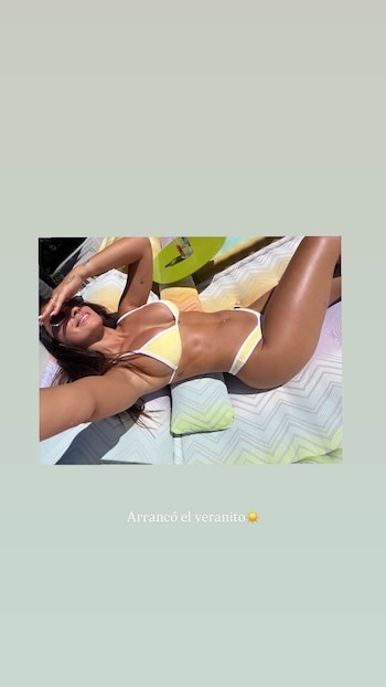 Antonela Roccuzzo con un bikini amarillo y blanco, gafas de sol y el cabello suelto, recostada en una tumbona a rayas bajo la luz del sol