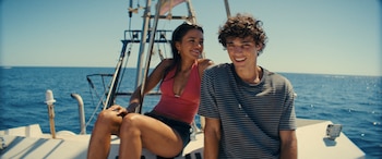 Dos jóvenes, una mujer y un hombre, sonríen sentados en la proa de un barco sobre el mar azul bajo un cielo despejado