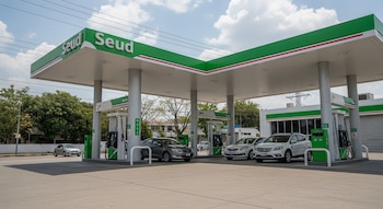 Estación de servicio Seud con dos marquesinas verdes y blancas, múltiples dispensadores de combustible y cuatro automóviles, bajo un cielo con nubes.