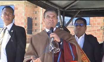 Primer plano del presidente Rodrigo Paz hablando por un micrófono, con poncho marrón. Flanqueado por dos hombres en traje, uno con gafas de sol. Fondo de ladrillos