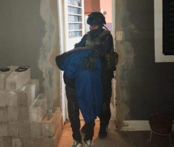 Persona con casco, chaleco y guantes tácticos carga a otra persona envuelta en tela azul, saliendo por una puerta. Hay bloques de hormigón