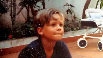 Fotografía de un niño de cabello claro en un patio exterior junto a un cochecito de bebé y plantas al fondo