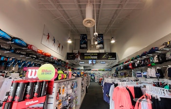 Pasillo central de una tienda deportiva con estanterías a ambos lados que exhiben raquetas, bolsos, pelotas de tenis, calzado e indumentaria