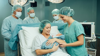 Madre en bata de hospital sostiene a su recién nacido envuelto en azul, sonriendo a una enfermera. Tres profesionales médicos con mascarillas están en el fondo.