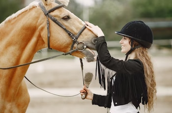 Primer plano de una mujer sonriente con casco negro y chaqueta con flecos acariciando el hocico de un caballo palomino con brida, sosteniendo un cepillo