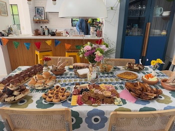 Mesa con mantel estampado, varios platos con medialunas, sándwiches, galletas, tabla de fiambres, ensalada, tarta, panes y arreglos florales