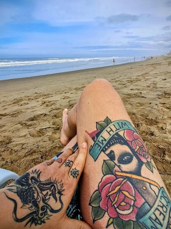 Piernas y una mano con tatuajes, incluyendo un dragón y un diseño de calavera con rosas, apoyadas en la arena de una playa con el mar de fondo