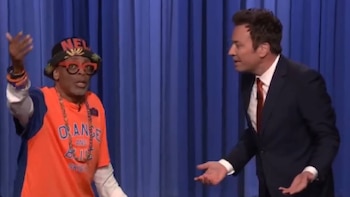 Dos hombres en un escenario azul. Uno, con camiseta naranja, gorro y gafas rojas, levanta el brazo. El otro, en traje oscuro, gesticula con ambas manos