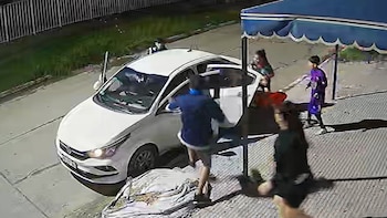 Vista aérea nocturna de varias personas alrededor de un auto blanco con puertas abiertas, frente a la entrada de un club con toldo azul en Aldo Bonzi