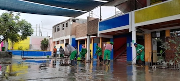 Siete personas con uniforme verde y botas de goma limpian un patio escolar anegado frente a edificios coloridos, árboles y una malla sombra