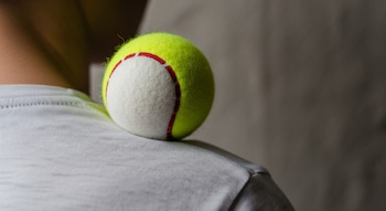 Primer plano de una pelota de tenis amarilla con líneas rojas apoyada en el hombro de una persona con camiseta gris. La persona está de pie contra una pared.
