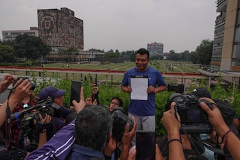 Un hombre sostiene un portapapeles con un documento mientras está rodeado por personas con cámaras fotográficas y teléfonos móviles frente a la Biblioteca Central de la UNAM